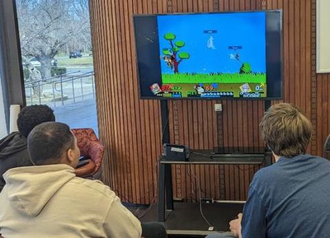 Three teen boys are playing Super Smash Bros on one of the Plainfield Public Library's big screens. The Smash Bros stage is based on the original Mario games.