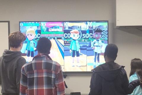 Four teenagers with their backs to the viewer are playing a lively game of Switch Sports Bowling on a big screen