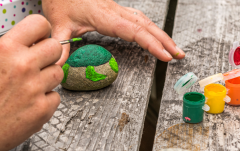 Hands shown painting a rock, apparently in a turtle design, over a wooden picnic bench surface. Small acrylic paint cups stand open on the right.