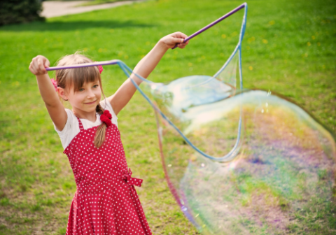 A tweenage girl in a red jumper and white shirt uses a string-and-stick bubble device to blow an enormous bubble against the background of a green field