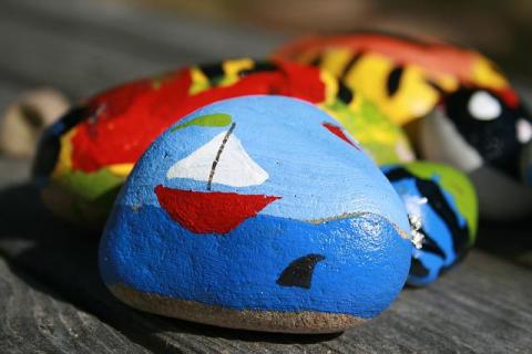 a selection of painted rocks, the front one being painted with a red sailboat with white sails on a blue sea