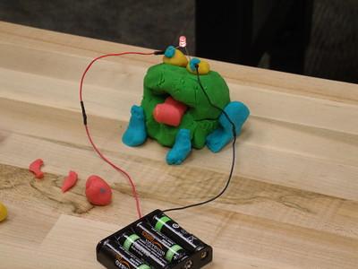 A multicolored frog made of playdough is connected by wires to a battery pack, making the LED lights in its eyes light up.