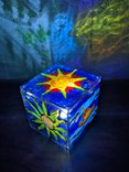 A small clear acrylic box painted with a star, blue background, and green patterns, is illuminated from within