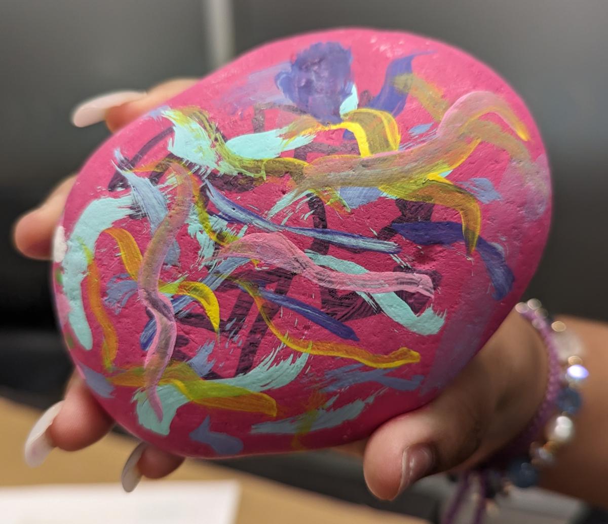 A hand holding a pink rock with multicolored waves over a figure reminiscent of Keith Haring