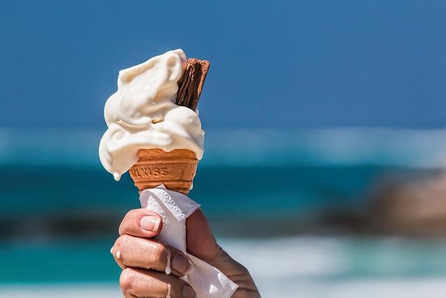 A hand holding a vanilla ice cream cone with a background of the beach